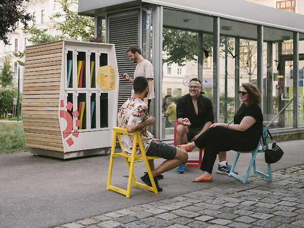 Foto eines Stadtmöbel-Regals und einer Gruppe Menschen, die auf Stühlen davor sitzt